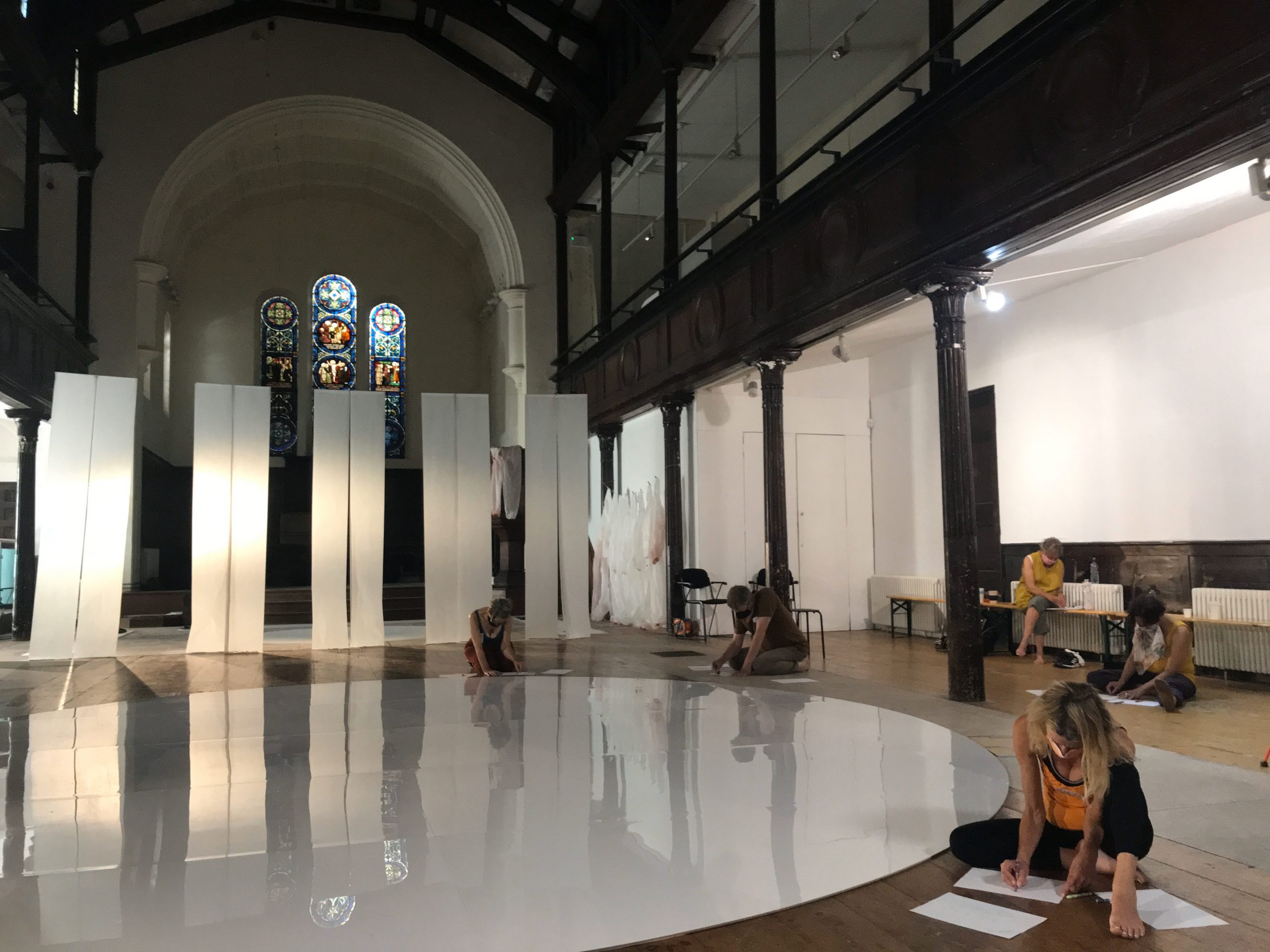 We can see five participant dancers, listening to soundscapes and responding in drawing, in the main gallery space at Fabrica, a former Georgian Church. We can see teh stained glass windows at the back
