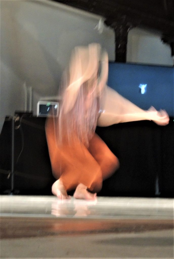 Blurred image of a dancer in movement on the dance floor, barefooted, with arms raised in the air, and orange trousers